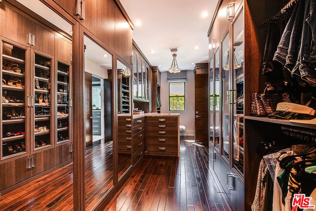 This is a luxurious walk-in closet featuring dark wood cabinetry with glass-fronted display cases for shoes and clothing. The closet includes built-in drawers and mirrored panels, enhancing the sense of space. The hardwood flooring adds warmth and elegance to the room.