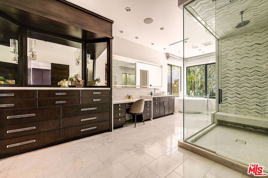 This is a luxurious primary bathroom featuring a large vanity with dark wood cabinetry and a white countertop, complemented by a spacious walk-in shower with patterned tile. A freestanding bathtub is visible through the glass shower enclosure, and the room is finished with marble flooring, creating a spa-like atmosphere. The perspective is from the entrance of the bathroom, showcasing the layout and high-end finishes.