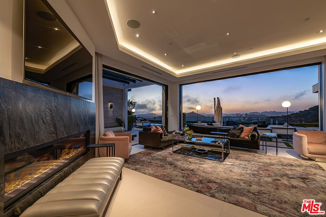 This is a luxurious living room featuring a modern fireplace with a large television above it. The room has floor-to-ceiling windows that offer a stunning view of the landscape at dusk. The furniture is stylish and comfortable, with a large area rug adding warmth to the space.