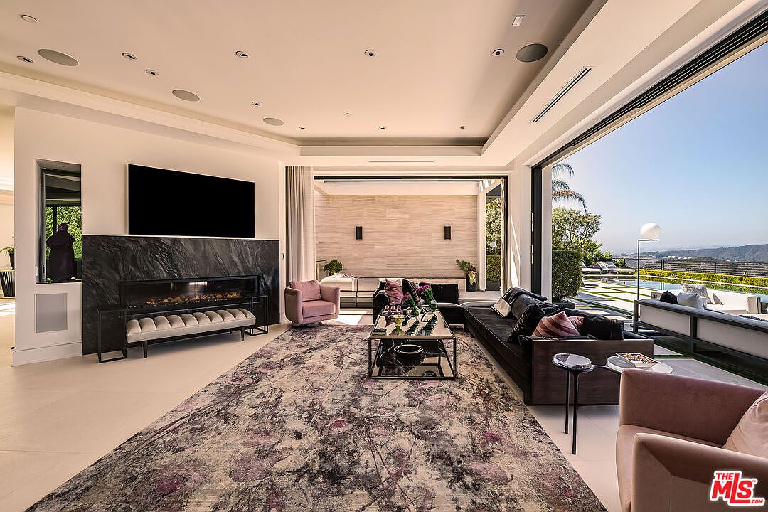 This is a luxurious living room featuring a modern fireplace with a large television above it, a plush area rug, and comfortable seating. The room opens to an outdoor patio area with a pool, offering stunning views. The style is contemporary and elegant, creating a sophisticated and inviting atmosphere.