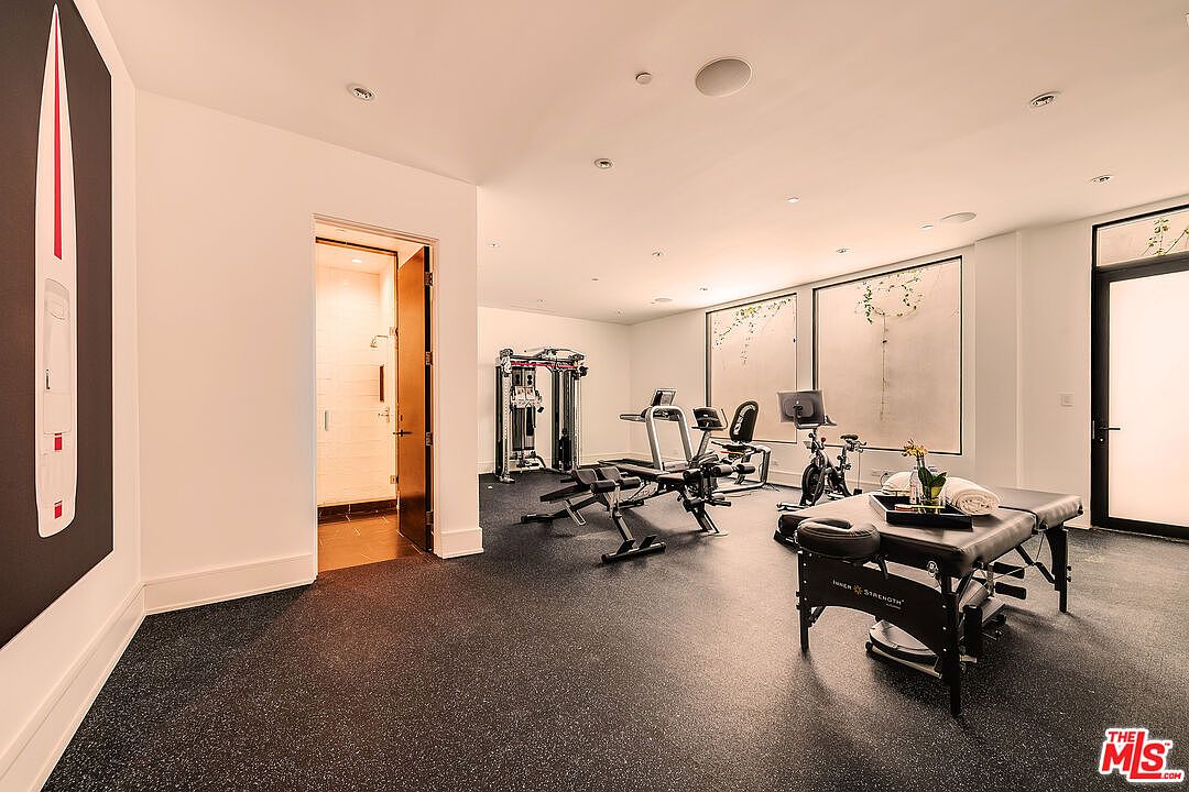 This is an interior shot of a home gym featuring various exercise machines, including a multi-station weight machine, stationary bikes, and a massage table. The room has a modern aesthetic with white walls, recessed lighting, and large windows that provide natural light. The flooring is a dark, rubberized material, and there is a doorway leading to what appears to be a bathroom or shower area.