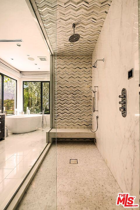 This is a luxurious primary bathroom featuring a spacious walk-in shower with chevron-patterned tile and a built-in bench. The shower is equipped with both a rainfall showerhead and a handheld sprayer. The bathroom also includes a freestanding bathtub visible through the glass partition, creating a spa-like atmosphere.