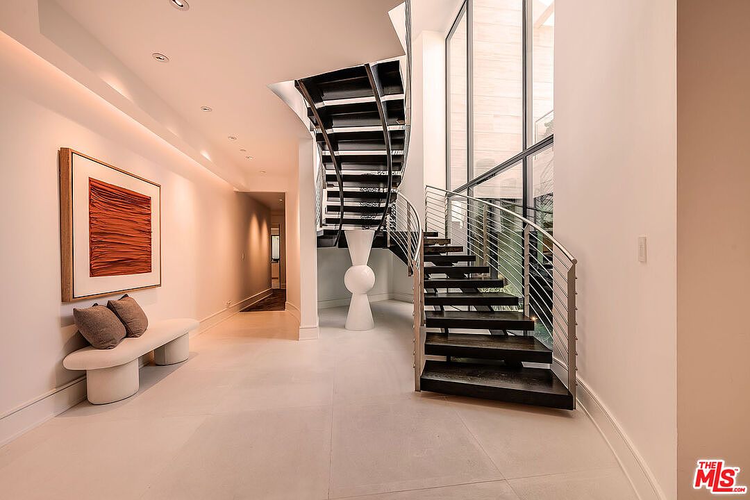 This interior shot showcases a modern hallway featuring a striking staircase with dark wood steps and a stainless steel railing. A large piece of artwork hangs on the wall above a minimalist bench with pillows, adding a touch of elegance. The space is well-lit with recessed lighting and natural light from the adjacent windows, creating a bright and inviting atmosphere.