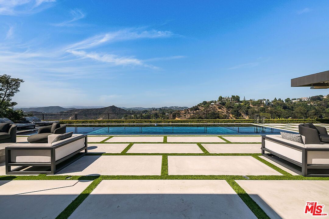 This image showcases a luxurious outdoor pool area with modern design elements. The pool is surrounded by a concrete patio with grass in between the concrete slabs, and stylish outdoor furniture is arranged on either side. The view extends to a scenic landscape with rolling hills and a clear blue sky, creating a serene and upscale ambiance.