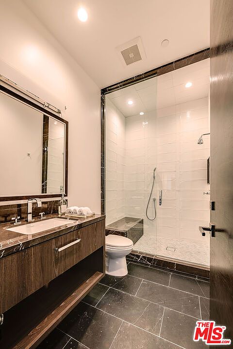 This is a modern primary bathroom featuring a dark wood vanity with a marble countertop and a large mirror. The shower is enclosed in glass with white tiled walls and a built-in bench. The flooring is dark tile, and the overall design is sleek and luxurious.