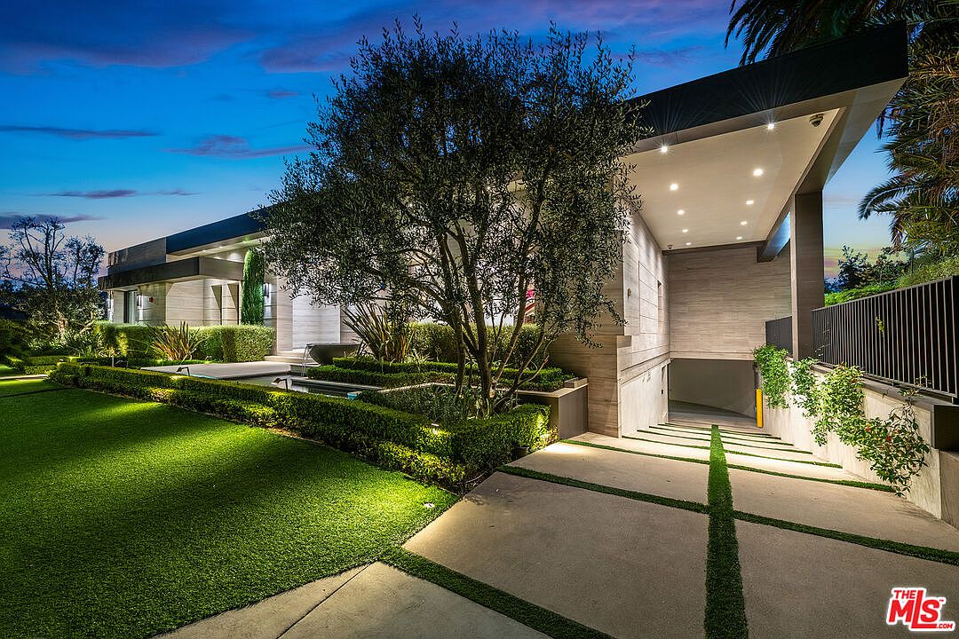 This image showcases the front exterior of a modern luxury home at dusk. The property features manicured lawns, sculpted hedges, and a striking architectural design with clean lines and a flat roof. A driveway leads to a subterranean garage, and the landscape is illuminated with strategic lighting, enhancing the property's curb appeal and highlighting its sophisticated aesthetic.