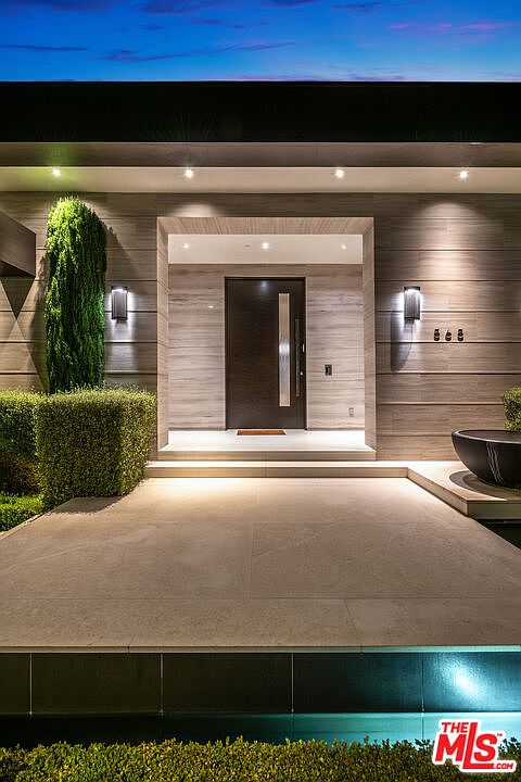 The image showcases a modern home's entryway, featuring a sleek, dark wood door framed by light-colored marble or stone. The entrance is flanked by manicured greenery and minimalist lighting fixtures, creating a sophisticated and inviting atmosphere. A shallow water feature adds a touch of elegance to the approach.
