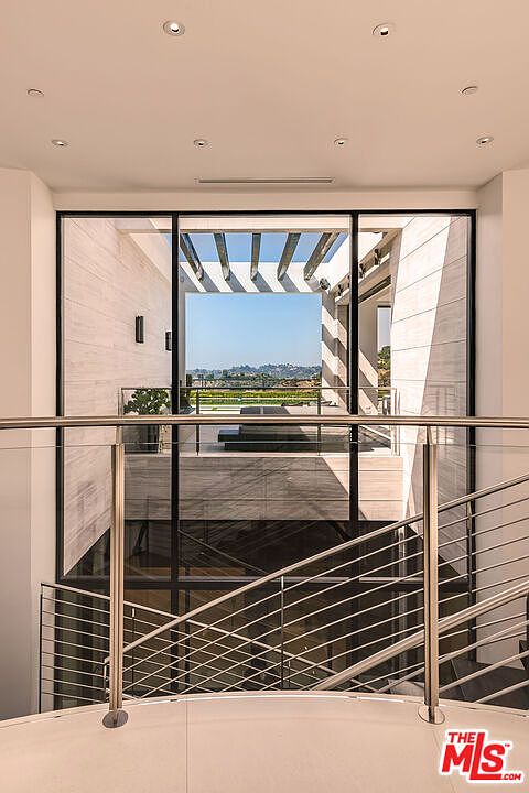 This interior shot showcases a modern hallway and staircase with sleek, stainless steel railings. A large window provides a view to the exterior, highlighting a patio or deck area with a pergola. The design emphasizes clean lines and a bright, airy atmosphere, creating a sophisticated and inviting space.