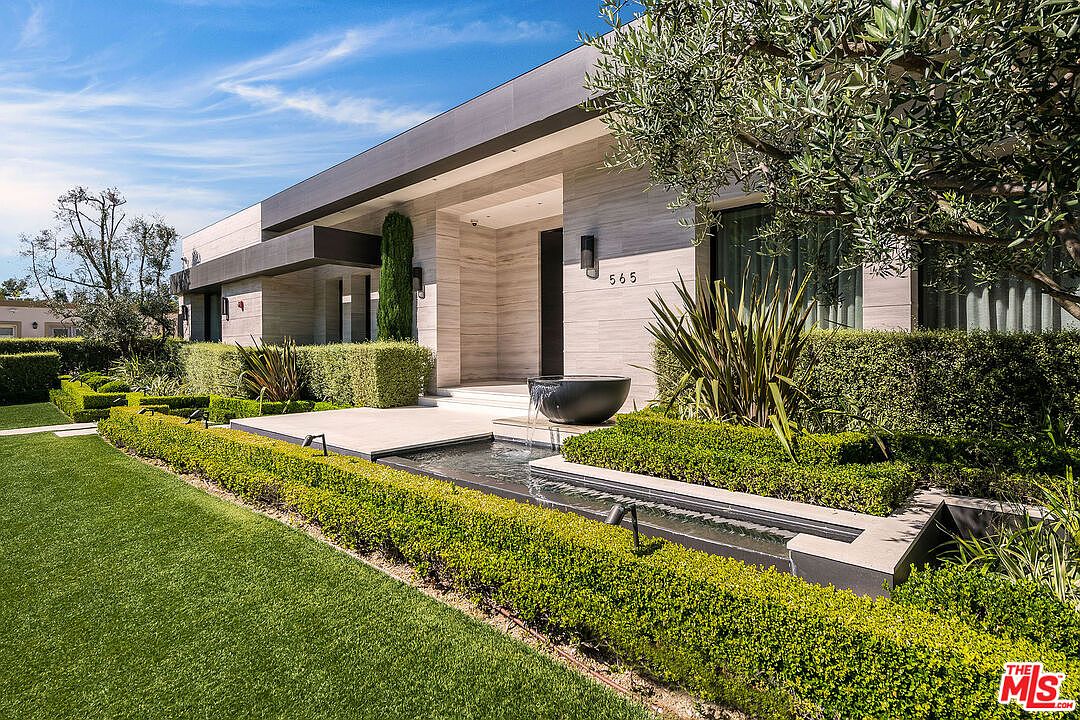 This is a front exterior view of a modern home. The house features a sleek, linear design with a light-colored facade and dark accents. A well-manicured lawn, hedges, and a water feature enhance the property's curb appeal, creating a sophisticated and inviting entrance.