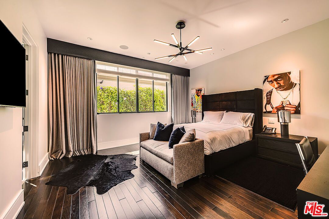 This is a stylish primary bedroom featuring dark hardwood floors, a large window with natural light, and a modern light fixture. The room includes a dark upholstered headboard, a small sofa at the foot of the bed, and artwork on the walls. The perspective is from the doorway, showcasing the room's layout and design elements.