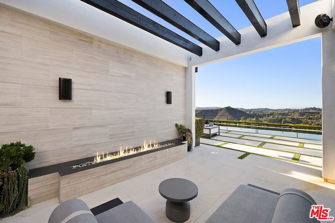 This image showcases a luxurious outdoor patio area with a modern design. A sleek linear fireplace is integrated into a tiled wall, complemented by contemporary outdoor furniture and a pergola overhead. The patio offers stunning views of the surrounding landscape, enhancing the property's appeal for relaxation and entertainment.