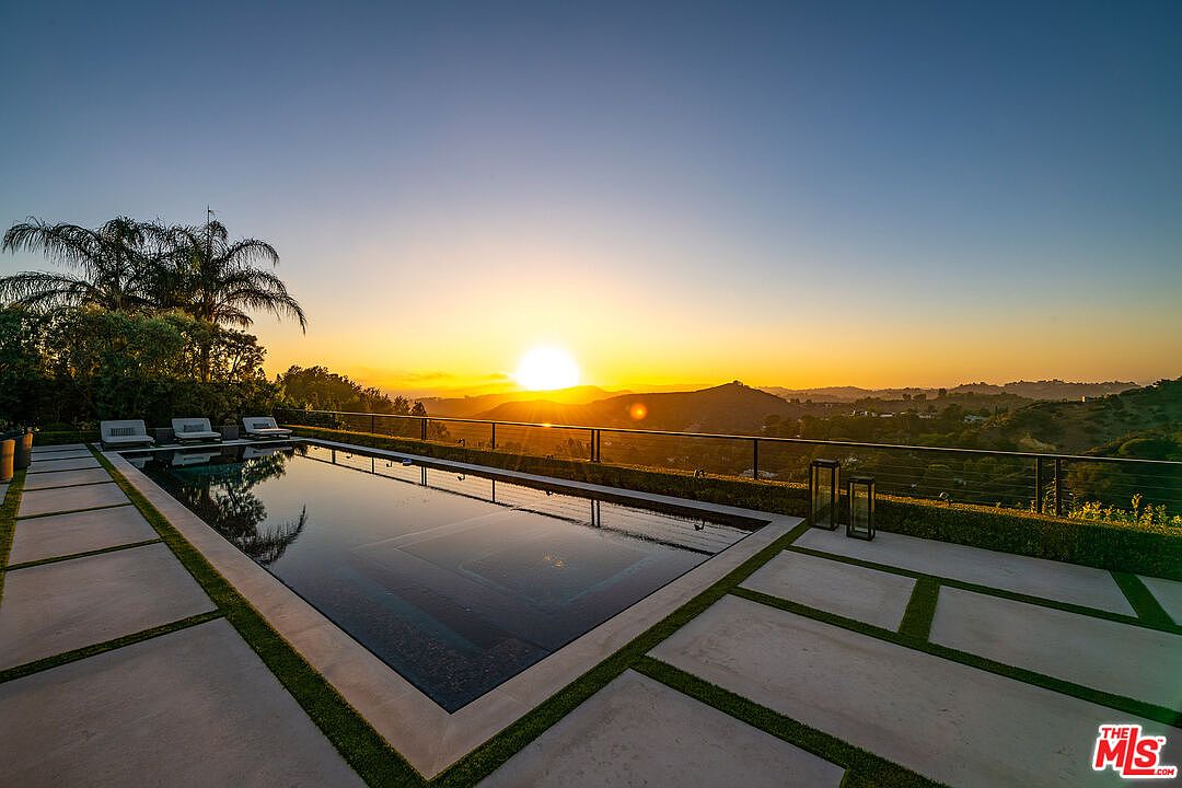 This image showcases a luxurious pool area at sunset, featuring a sleek, modern pool with dark water reflecting the sky. The pool is surrounded by a concrete patio with grass inlays, and lounge chairs are placed at one end. A metal railing provides safety and unobstructed views of the distant hills and vibrant sunset, creating a serene and upscale ambiance.