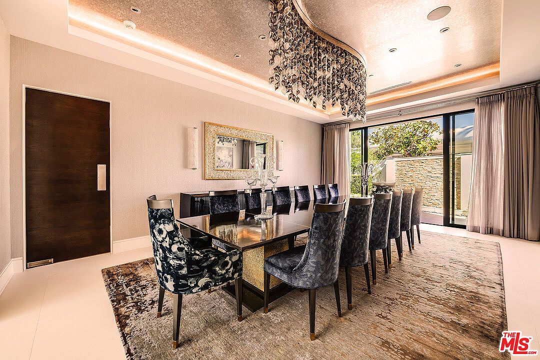 This is an interior shot of a luxurious dining room. The room features a long, dark wood dining table with gold accents, surrounded by upholstered chairs with unique patterns. A large, ornate chandelier hangs above the table, and a mirror adorns the wall, reflecting the light and space. The room opens to an outdoor area through large glass doors, creating a seamless transition between indoor and outdoor living.