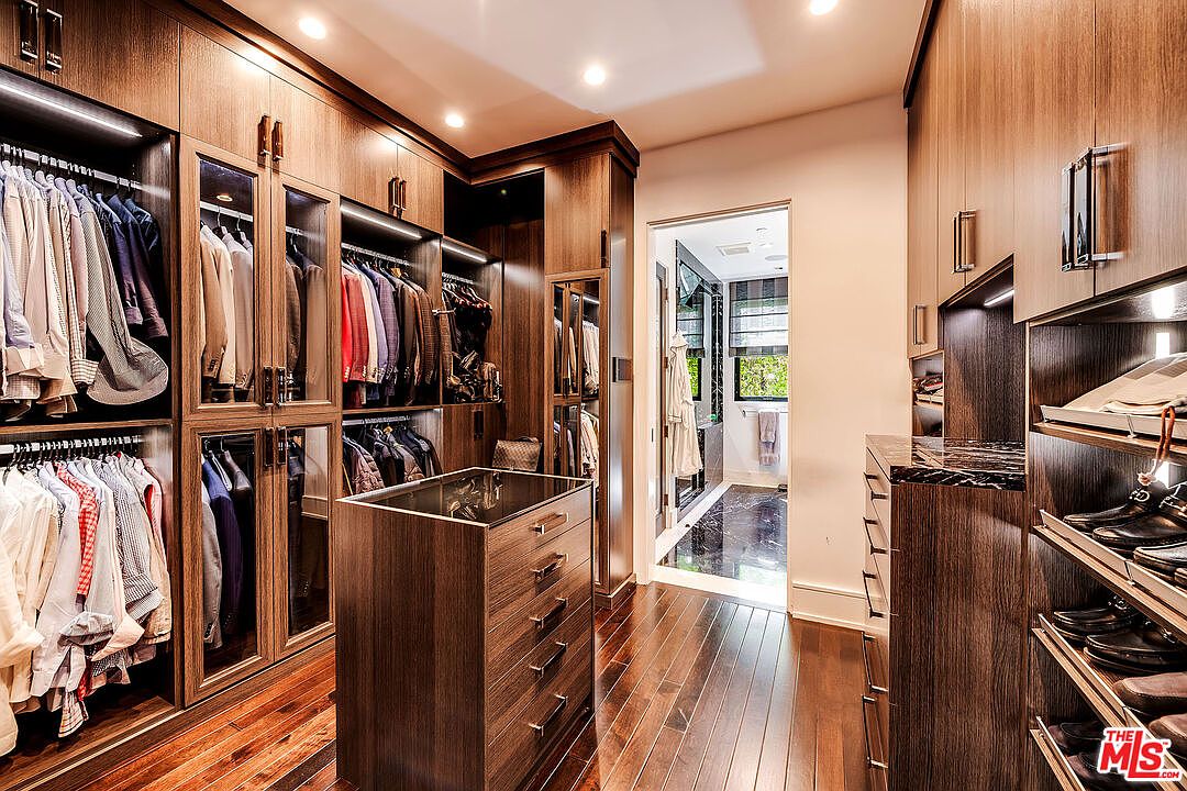 This is a luxurious walk-in closet featuring custom wood cabinetry with glass-fronted doors and integrated lighting. A central island with drawers provides additional storage, and hardwood flooring adds warmth to the space. The closet leads to a bathroom, suggesting a primary suite.