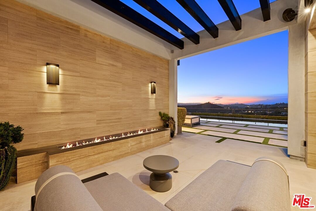 This image showcases a luxurious outdoor patio area with a modern design. The space features a built-in fireplace, sleek lounge chairs, and a small side table, all set against a backdrop of a stunning sunset view. The patio's design incorporates clean lines and natural materials, creating a sophisticated and relaxing atmosphere.