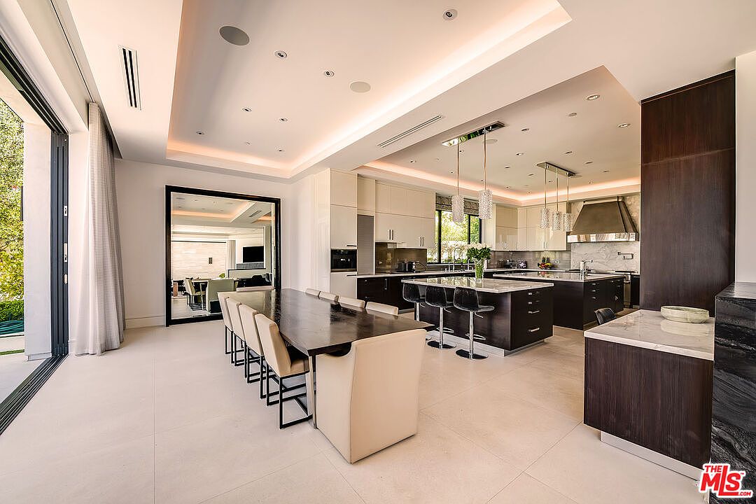 This is a spacious, modern kitchen and dining area featuring a large dining table with seating for ten, a sleek kitchen island with bar stools, and high-end appliances. The kitchen boasts a combination of light and dark cabinetry, marble countertops, and pendant lighting, creating a luxurious and functional space. Large sliding glass doors provide ample natural light and access to the outdoors.