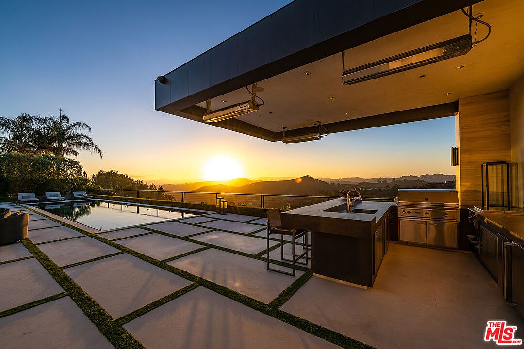 This image showcases a luxurious outdoor patio area at sunset, featuring a modern design with a pool, a built-in outdoor kitchen with stainless steel appliances, and a covered seating area. The patio is paved with large square tiles interspersed with grass, offering a clean and contemporary aesthetic. The view overlooks a scenic landscape of rolling hills and a vibrant sunset, enhancing the property's appeal.