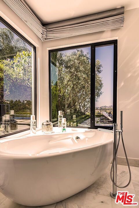 This is a luxurious primary bathroom featuring a freestanding white bathtub positioned in front of a large window with a view of lush greenery. The bathroom is well-lit, showcasing a modern design with clean lines and a minimalist aesthetic. A sleek, chrome faucet stands beside the tub, enhancing the room's sophisticated ambiance.