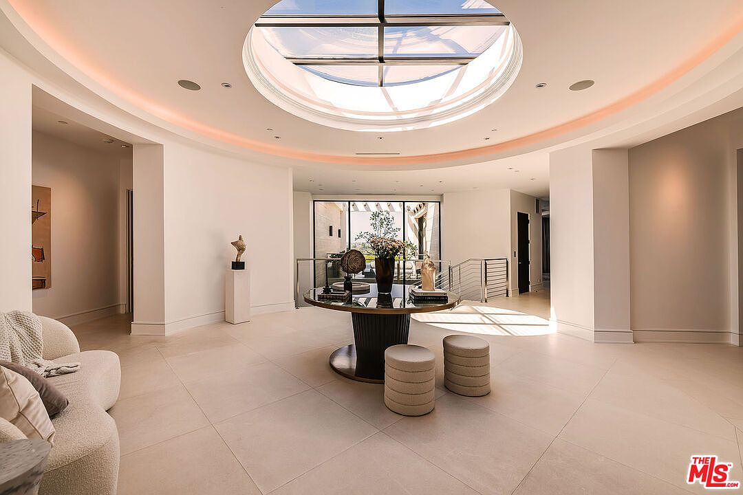 This interior shot showcases a grand hallway with a striking circular skylight, illuminating the space with natural light. A round table with decorative items and two ottomans anchor the center, while a glimpse of the staircase and a seating area add depth. The neutral color palette and clean lines contribute to a luxurious and modern aesthetic.