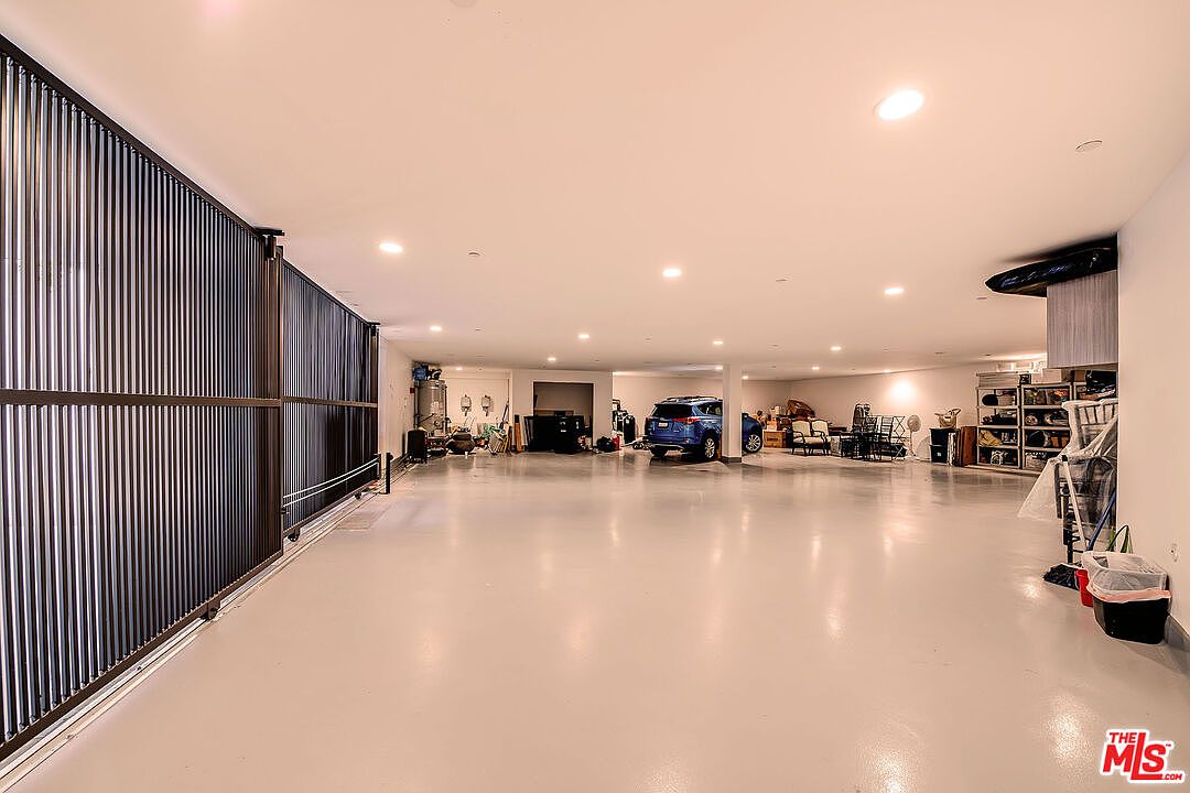 This is a spacious basement featuring a large open area with a light gray floor and white walls. A metal slat wall is on the left, and a blue SUV is parked in the background. The room is well-lit with recessed lighting, and there are various items stored along the back wall, suggesting ample storage space.