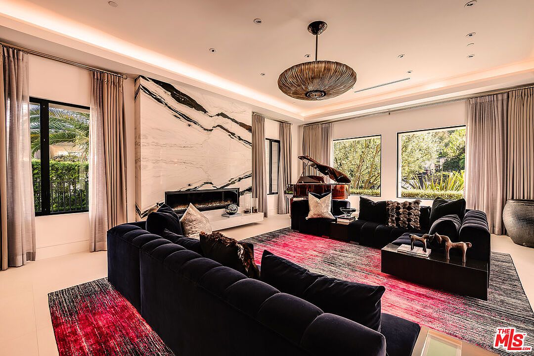This is a luxurious living room featuring a modern fireplace with a marble surround, a grand piano, and plush, dark-colored sofas. The room is illuminated by a unique pendant light fixture and natural light streaming through large windows, creating a sophisticated and inviting atmosphere. A patterned rug adds a touch of color and texture to the space.