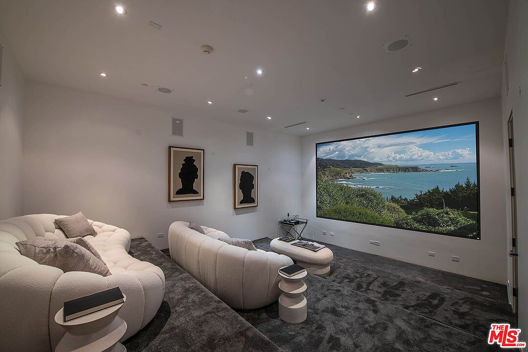 This is a modern living room featuring two curved white sofas, gray carpeting, and two framed silhouette artworks on the wall. A large screen displays a coastal landscape, creating a focal point. The room is well-lit with recessed lighting, contributing to a clean and minimalist aesthetic.