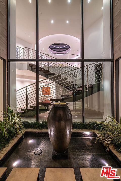 This interior shot showcases a modern staircase with sleek metal railings and wooden steps, visible through a large glass window. A decorative water feature with a large vase sits in the foreground, adding a tranquil element to the space. The overall design is contemporary and emphasizes natural light and architectural details.