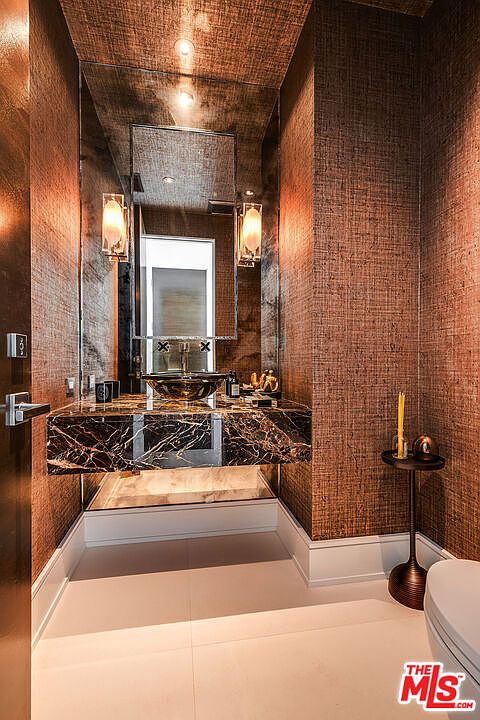 This is a stylish guest bathroom featuring a unique marble countertop with a vessel sink and modern fixtures. The walls are covered in a textured, metallic-toned wallpaper, complemented by sconce lighting and a large mirror that enhances the sense of space. The overall design is luxurious and contemporary, creating a sophisticated powder room.
