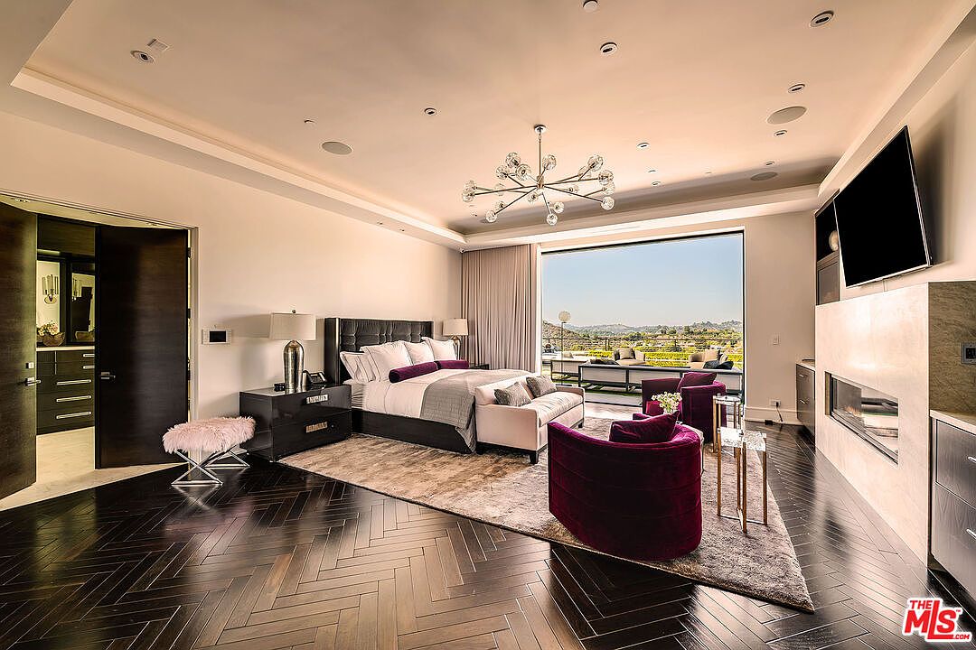 This is a luxurious primary bedroom featuring dark hardwood floors in a herringbone pattern, a large bed with a tufted headboard, and a seating area with plush armchairs. A large window offers a view of the landscape, and a modern chandelier hangs from the ceiling. The room exudes elegance and comfort.