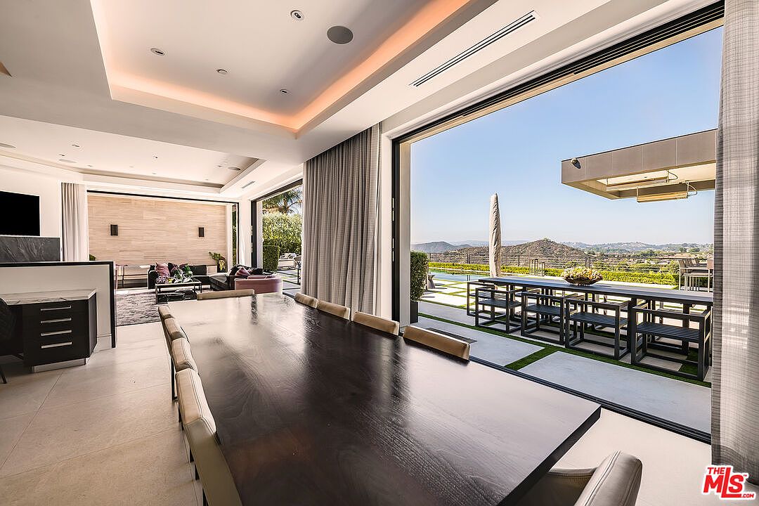 This interior shot showcases a modern dining room with a long, dark wood table and light-colored chairs. Large sliding glass doors open to an outdoor patio with a dining area and views of the landscape. The room features recessed lighting and neutral tones, creating a sophisticated and inviting atmosphere.