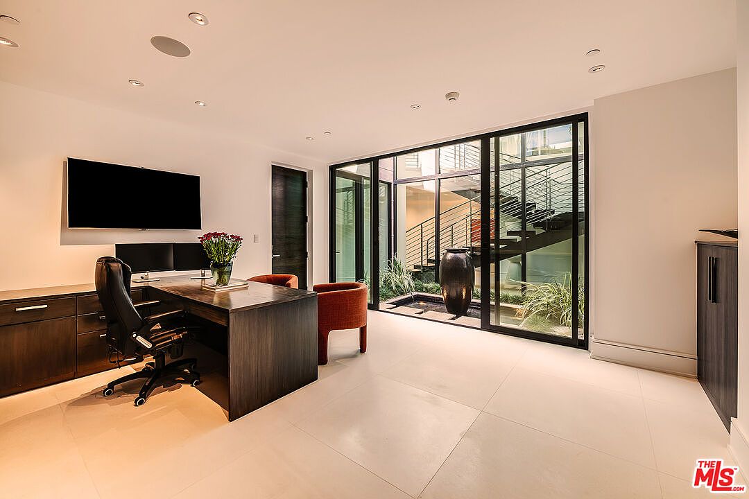 This is an interior shot of a modern office space featuring a dark wood desk with a black leather office chair, a large flat-screen TV mounted on the wall, and a floor-to-ceiling window offering a view of an outdoor courtyard with stairs. The room is well-lit with recessed lighting and features neutral-toned tile flooring, creating a clean and professional atmosphere.