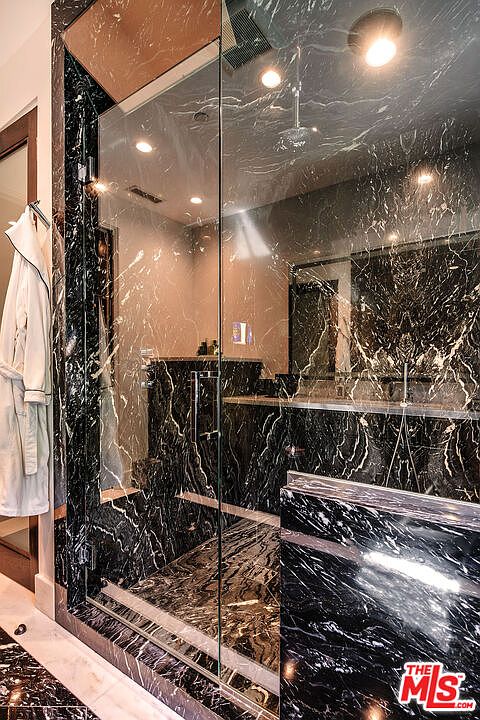 This is a luxurious primary bathroom featuring black marble walls and flooring with striking white veining. A glass-enclosed shower is the focal point, showcasing the continuation of the marble design within. A white robe hangs on the wall, adding a touch of elegance to the space.