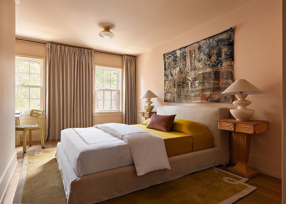 This elegant primary bedroom features a neutral color palette with warm, earthy tones, centered around a plush upholstered bed dressed in crisp white linens and a mustard-yellow accent layer. A large, textured tapestry hangs above the headboard, flanked by unique sculptural table lamps on wooden nightstands, while floor-to-ceiling curtains frame the windows. The room is anchored by a soft, mustard-colored area rug, creating a cozy and sophisticated atmosphere.