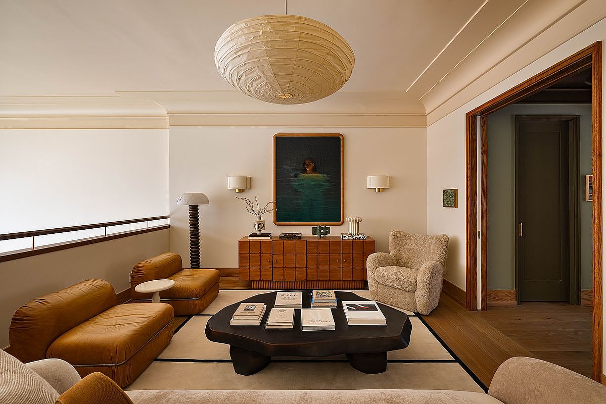 This sophisticated living room features a warm, curated aesthetic with a mix of plush, textured seating and a striking, organic-shaped dark wood coffee table. A large, textured paper pendant light serves as a focal point above the seating area, while a framed portrait hangs above a mid-century modern wooden console. The space is defined by a neutral color palette, clean architectural lines, and a sense of refined, quiet luxury.