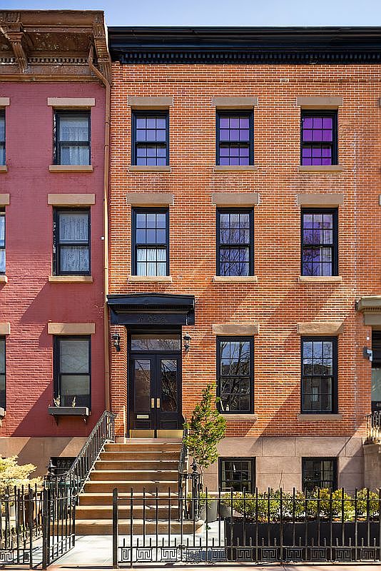 This image captures a classic, multi-story brick townhouse from a straight-on, eye-level perspective, showcasing its traditional architectural charm. The facade features symmetrical window placement, a prominent dark-framed entryway with a stoop, and a decorative cornice at the roofline. A wrought-iron fence and a small, well-maintained garden bed in the foreground add to the inviting, urban residential aesthetic.