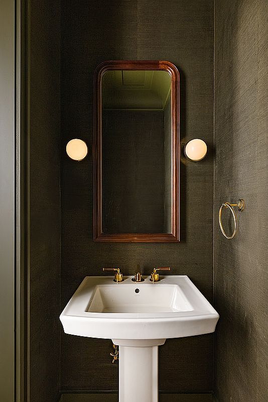 This elegant powder room features a classic pedestal sink with polished brass fixtures set against a dark, textured wallpaper that creates a sophisticated and moody atmosphere. A rectangular mirror with a warm wood frame is flanked by two minimalist globe wall sconces, providing soft, ambient lighting. The composition is centered and symmetrical, emphasizing a refined, high-end design aesthetic.