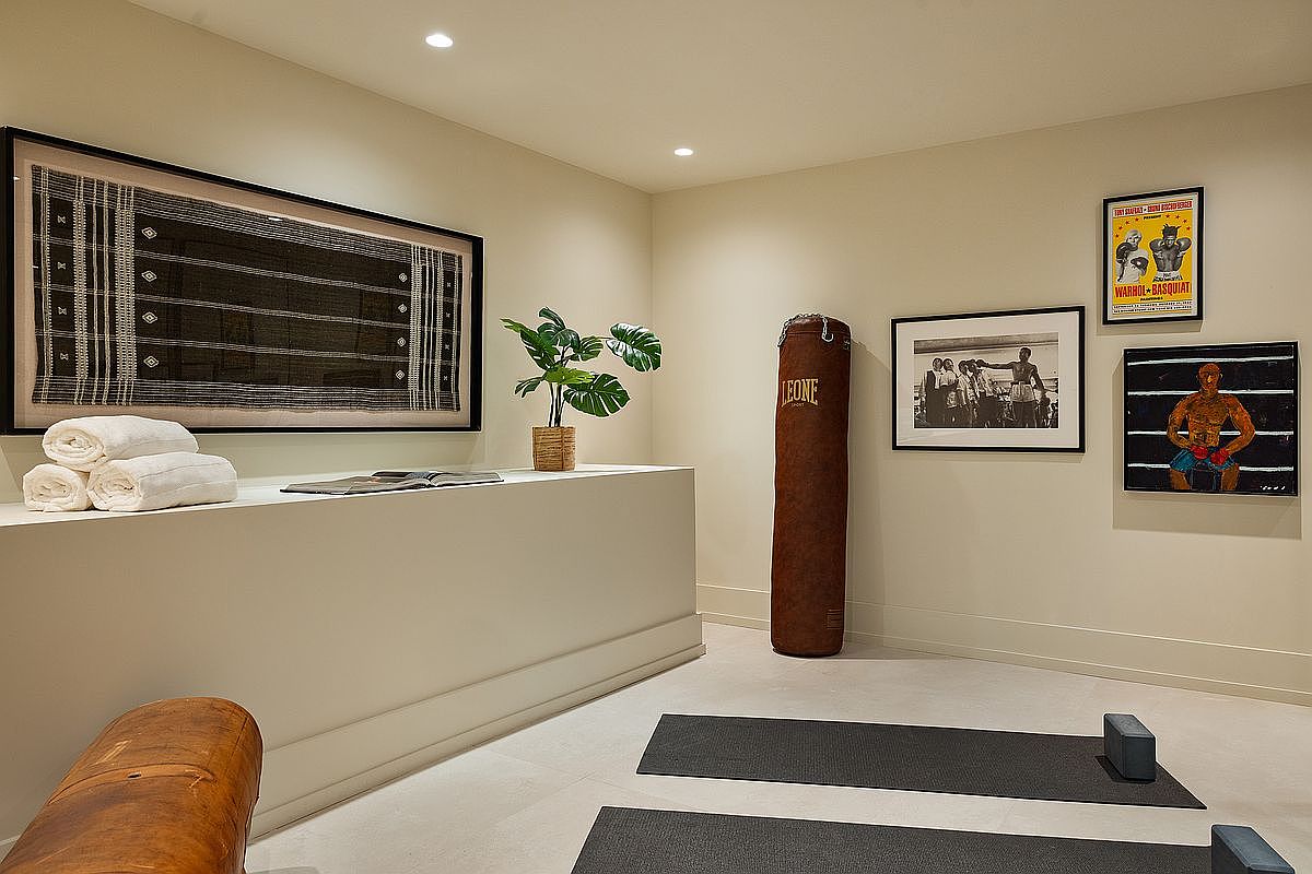 This sophisticated home gym features a minimalist aesthetic with neutral walls, a brown leather punching bag, and two yoga mats laid out on the floor. The space is accented with curated wall art, including a boxing-themed poster and a framed textile piece, creating a serene yet focused workout environment. The perspective is eye-level, capturing the clean lines and balanced layout of this dedicated fitness area.