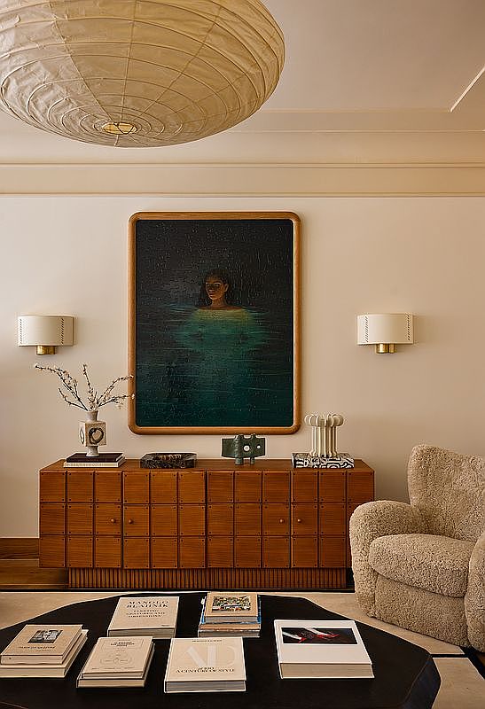 This sophisticated living room space features a warm, textured aesthetic centered around a large, dark-toned painting of a figure in water, flanked by elegant wall sconces. Below the artwork sits a unique, grid-patterned wooden credenza, while a plush, cream-colored bouclé armchair and a dark, organic-shaped coffee table topped with curated books complete the cozy seating area. The room is illuminated by a large, sculptural paper pendant light, creating a serene and high-end atmosphere.
