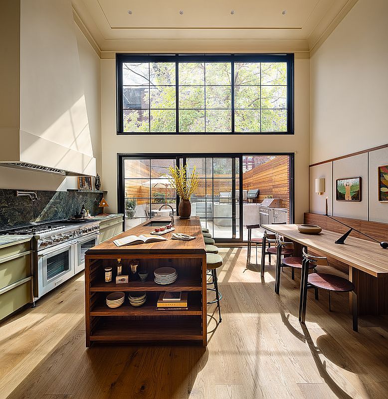 This sophisticated, high-ceilinged kitchen features a large wooden island with integrated shelving and a seamless transition to an adjacent dining area. The space is anchored by a professional-grade range, dark stone backsplash, and expansive floor-to-ceiling windows that flood the room with natural light while providing a view of the private patio. The warm wood tones and clean, modern lines create an inviting and luxurious atmosphere perfect for both cooking and entertaining.