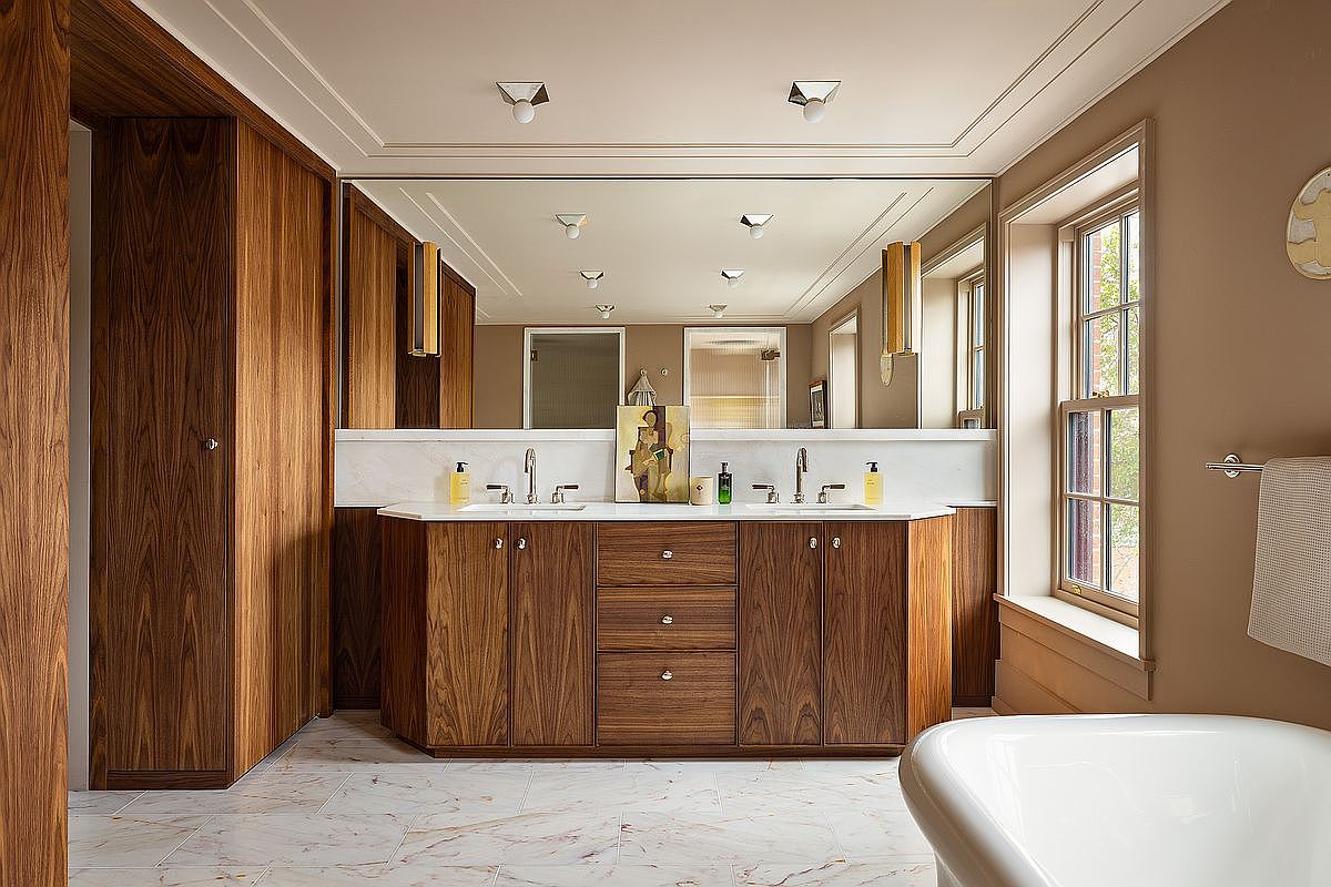 This elegant primary bathroom features a sophisticated mid-century modern aesthetic with rich, warm-toned walnut cabinetry and a clean white marble countertop. The space is anchored by a large, seamless wall-to-wall mirror that reflects the room's recessed lighting and a freestanding soaking tub visible in the foreground. The combination of natural wood textures, marble flooring, and soft neutral wall tones creates a luxurious and serene atmosphere.