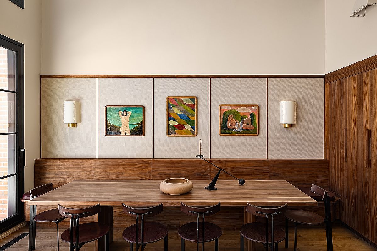 This sophisticated dining room features a warm, mid-century modern aesthetic with a rich walnut wood dining table and matching curved chairs. The wall is elegantly paneled with fabric-upholstered sections, adorned with three framed artworks and minimalist wall sconces, creating a gallery-like atmosphere. A sleek, sculptural candle holder and a ceramic bowl serve as refined centerpieces on the table, while the overall composition exudes a sense of curated luxury and calm.