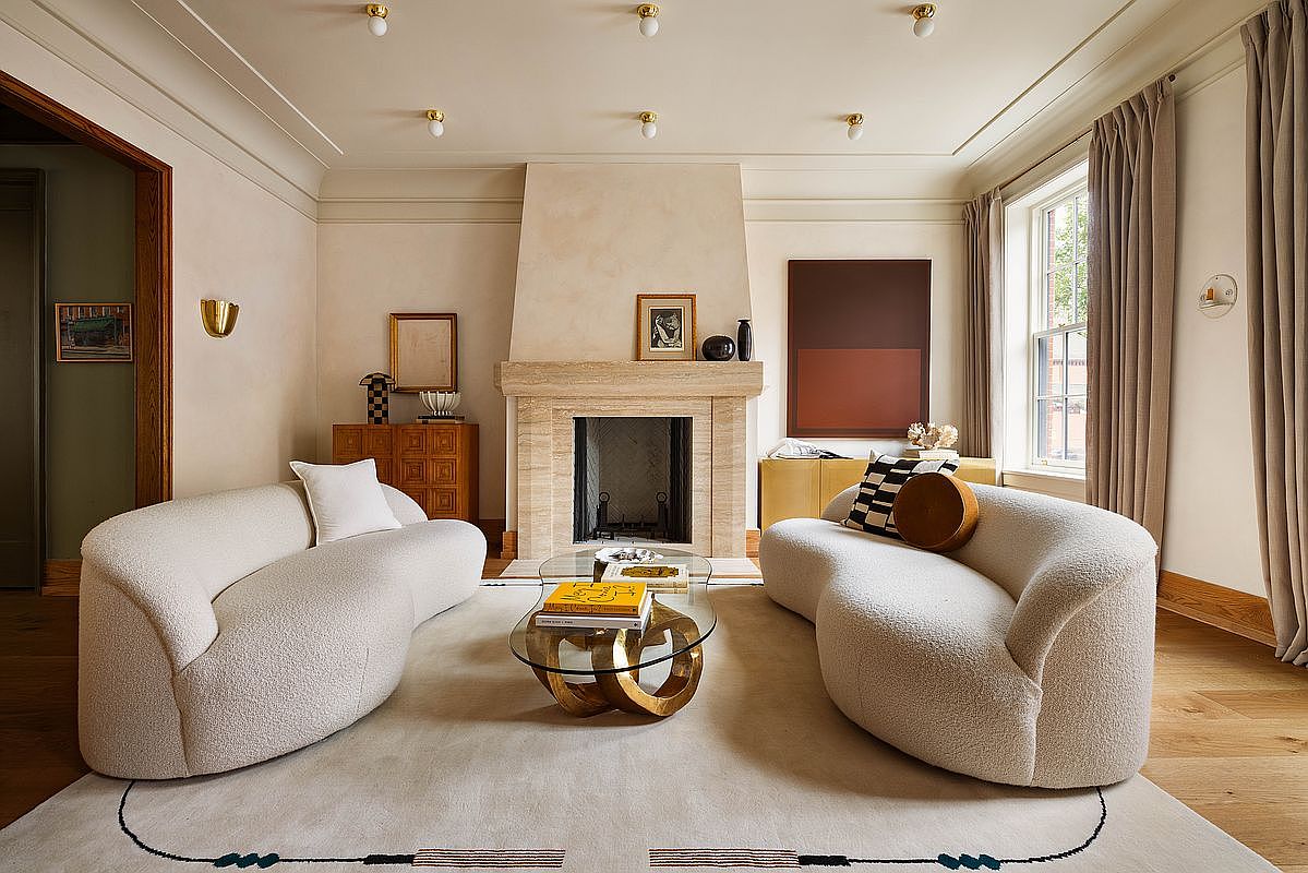 This sophisticated living room features a symmetrical layout with two curved, cream-colored bouclé sofas facing each other across a sculptural gold-based coffee table. A prominent travertine fireplace serves as the central focal point, flanked by minimalist art and warm, neutral wall tones. The space is grounded by a large area rug with subtle linear details, creating an inviting and high-end atmosphere perfect for entertaining.