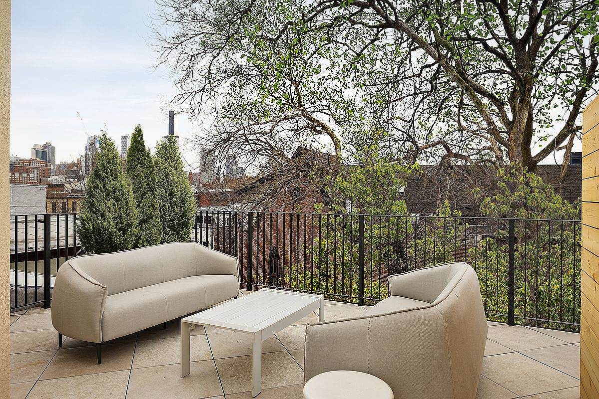 This modern rooftop terrace features a sleek, minimalist seating area with a light-colored sofa and matching armchair arranged around a white coffee table. The space is framed by a black metal railing and offers a scenic view of the surrounding neighborhood trees and distant city skyline. The paved stone flooring and clean lines create an inviting, sophisticated outdoor living environment perfect for relaxation or entertaining.
