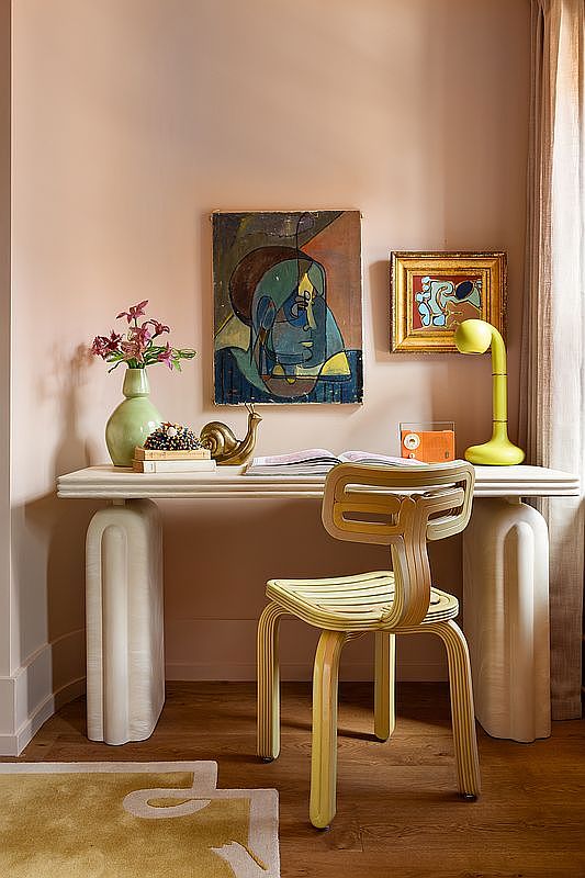 This stylish home office nook features a unique, sculptural desk with rounded, textured legs and a matching bentwood chair in a soft yellow hue. The space is accented by a vibrant abstract painting, a modern lime-green desk lamp, and a delicate floral arrangement, all set against a warm, peach-toned wall. The composition offers a cozy, artistic atmosphere perfect for creative work.