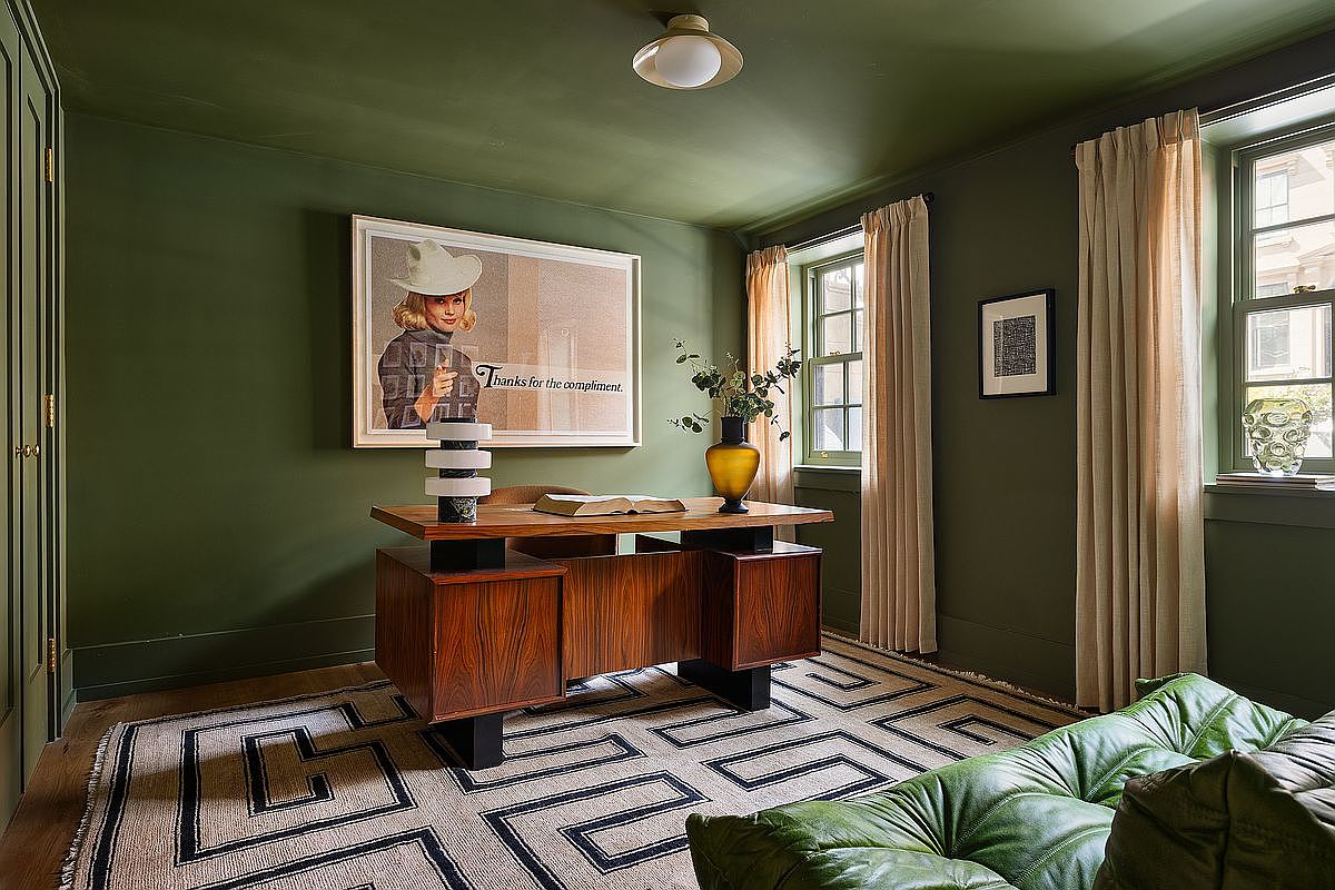 This sophisticated home office features deep olive-green walls and ceiling, creating a moody and focused atmosphere. A mid-century modern wooden desk serves as the centerpiece, complemented by a geometric patterned rug and a plush green leather sofa in the foreground. The room is accented by a large framed art piece, elegant beige curtains, and warm ambient lighting, offering a refined and professional aesthetic.