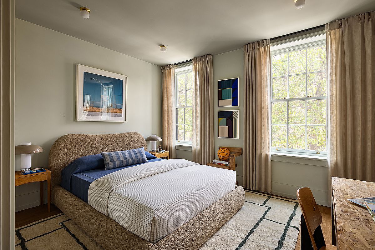 This contemporary bedroom features a textured, rounded-edge bed frame as the centerpiece, complemented by a blue accent pillow and a neutral-toned grid-patterned duvet. The room is bathed in natural light from two large windows dressed in floor-to-ceiling beige curtains, while modern art pieces and a unique wooden chair add a curated, artistic flair. The overall aesthetic is warm, minimalist, and inviting, with a soft area rug grounding the space.