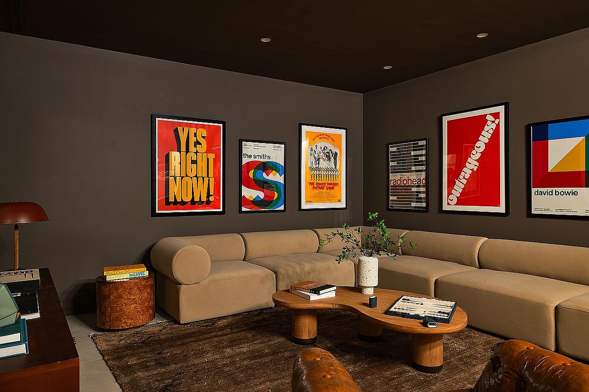 This stylish, moody living room features dark charcoal walls adorned with a curated collection of vibrant music and pop culture posters. A large, plush beige sectional sofa anchors the space, complemented by a unique, organic-shaped wooden coffee table and a rich brown area rug. The room exudes a sophisticated, eclectic vibe, perfect for relaxation or entertainment.