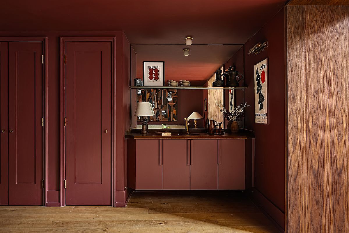 This sophisticated powder room features a bold, monochromatic deep burgundy color palette that extends from the walls to the cabinetry and ceiling. A large, wall-to-wall mirror reflects the space, highlighting a sleek vanity with a dark countertop, a minimalist lamp, and curated art pieces. The warm wood flooring and rich textures create a moody, high-end atmosphere that feels both intimate and luxurious.