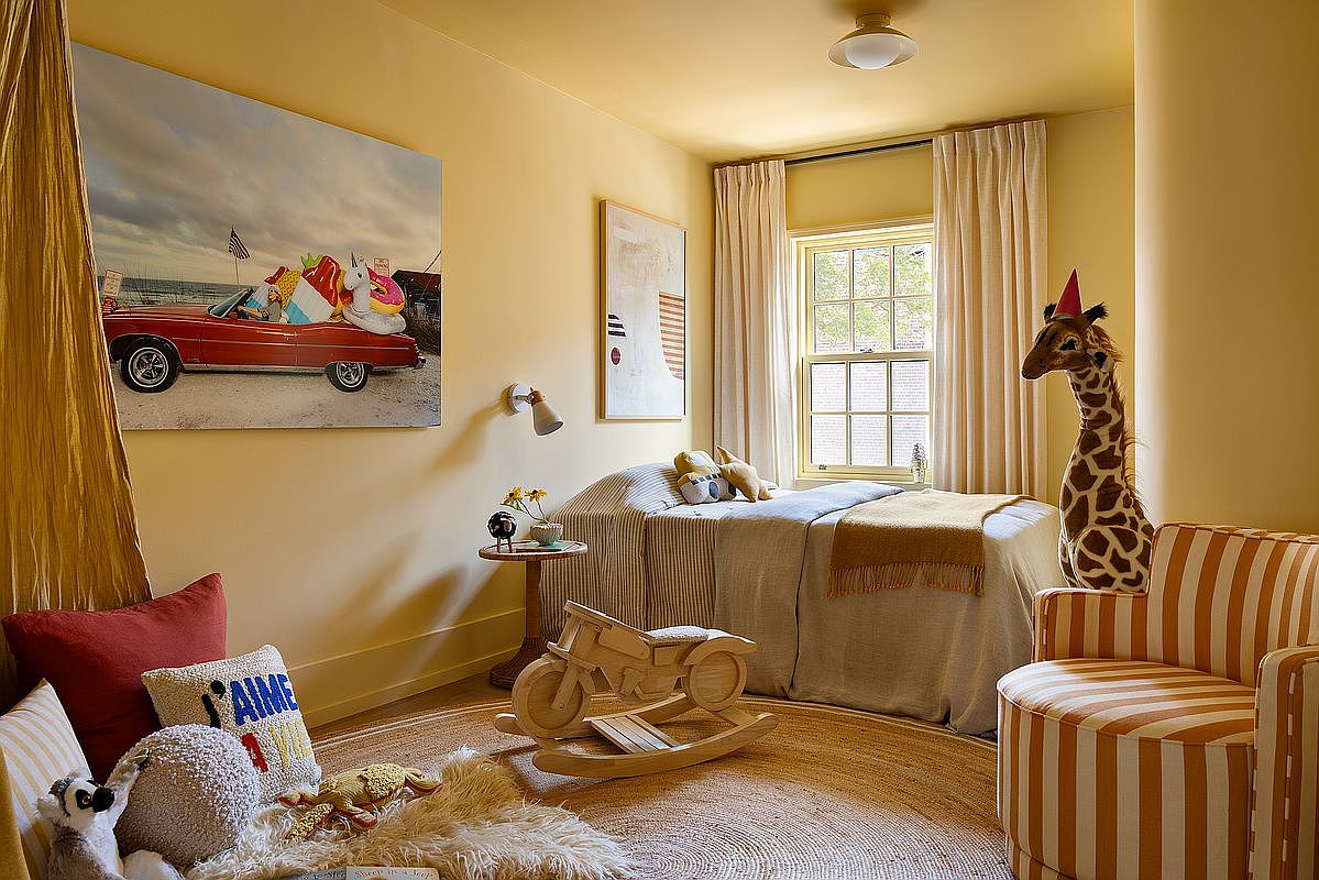 This charming, sun-drenched child's bedroom features a warm yellow color palette, a cozy twin bed with neutral linens, and a whimsical giraffe plush toy wearing a party hat. The room is decorated with playful elements like a wooden rocking motorcycle, a large beach-themed wall art piece, and a striped armchair, creating a cheerful and inviting atmosphere. The perspective is a wide, eye-level shot that captures the entire room's layout and its delightful, curated decor.