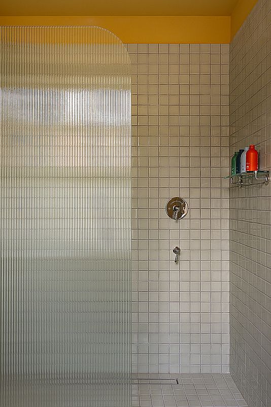 This minimalist shower space features a distinctive curved, fluted glass partition that adds a modern, textured aesthetic. The walls are clad in small, square white tiles, complemented by a vibrant yellow accent wall at the top, while a sleek chrome shower fixture and a glass shelf with colorful toiletries complete the functional design.